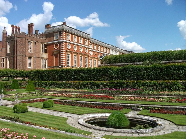 Gardens of Hampton Court in England with manicured hedges, colorful flowerbeds, and decorative fountains set against the red-brick Tudor and Baroque palace buildings under a bright blue sky, showcasing the grandeur of the royal estate