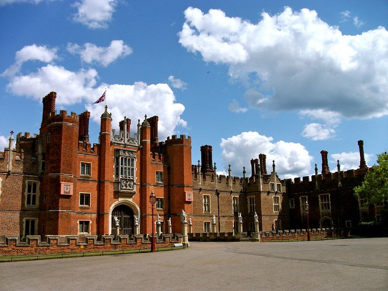 Exterior view of Hampton Court Palace in England featuring red-brick Tudor architecture, tall chimneys, arched gateway, and historic towers under a bright blue sky with clouds, highlighting the grandeur of this former royal residence