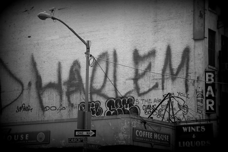 Black and white photo of a Harlem graffiti mural painted across a brick wall above storefronts in New York City, including signs for a coffee house and a liquor store. The gritty urban streetscape captures the neighborhood’s distinctive character and vibe