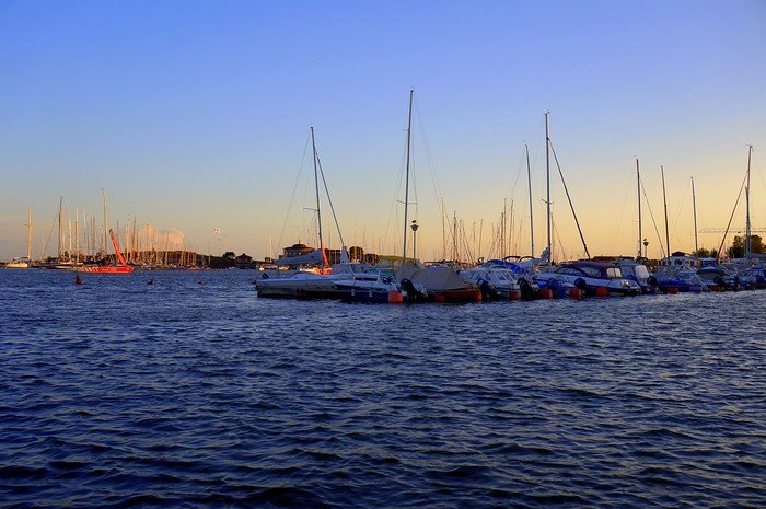 Sunset over the harbour in Helsinki, Finland, with sailboats and yachts docked in the marina, calm waters reflecting the golden light of evening, and the sky transitioning from warm tones to deep blue at the close of day