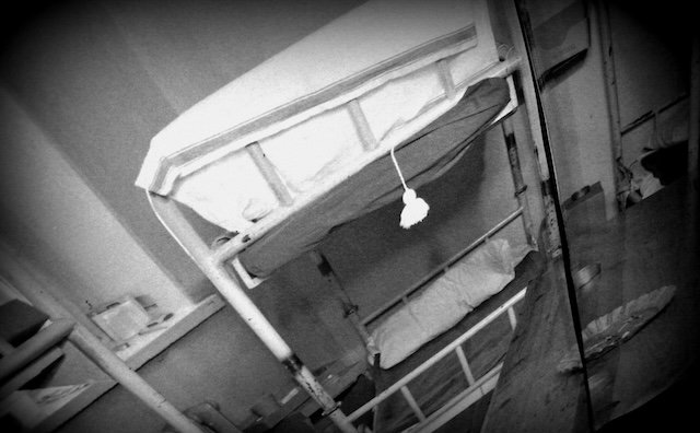 Black and white retro photo of an old prison cell at Hotel Katajanokka in Helsinki, Finland, showing metal bunk beds, simple bedding, and stark furnishings. The historic image reflects the building’s past life as a functioning jail before its hotel conversion