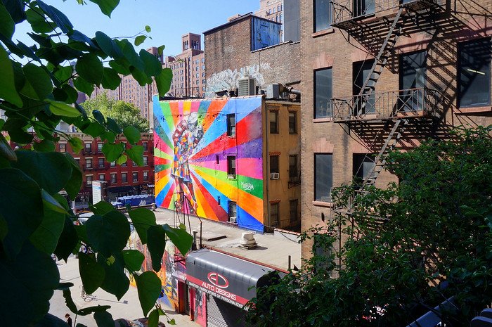 View of a vibrant rainbow-colored mural depicting a famous kiss, seen from the High Line in New York City on a sunny summer day. The artwork stands out against brick buildings with fire escapes, leafy green plants in the foreground, and clear blue skies