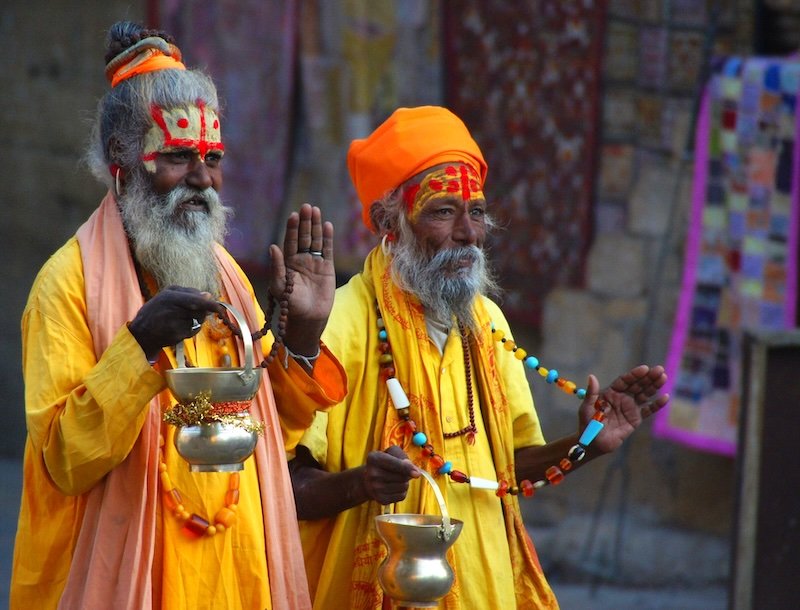 Two Hindu holy men in Jaisalmer, Rajasthan, India wearing bright saffron robes, painted foreheads, and beaded necklaces while holding brass offering pots and blessing passersby