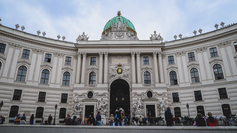 The grand Baroque façade of Hofburg Palace in Vienna, Austria, with its ornate sculptures, arched entrance, and iconic green dome, bustling with visitors in the courtyard The grand Baroque façade of Hofburg Palace in Vienna, Austria, with its ornate sculptures, arched entrance, and iconic green dome, bustling with visitors in the courtyard