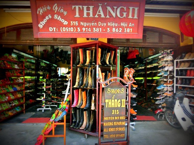 Traditional shoe shop in Hoi An, Vietnam, with rows of colorful handmade leather shoes and tall boots displayed on racks at the entrance, a wooden sign advertising custom shoe making and repairs, and vibrant shelves of footwear lining the interior of the yellow-painted storefront