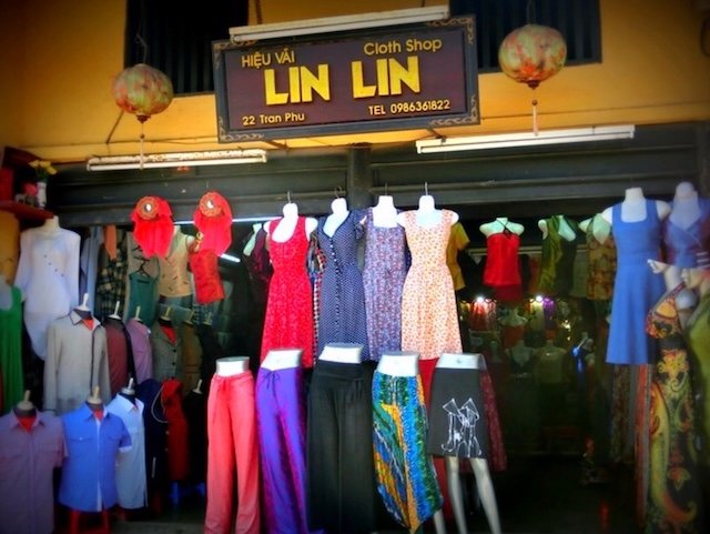 Exterior of Lin Lin Cloth Shop in Hoi An, Vietnam, with an array of colorful custom-made dresses, skirts, pants, and shirts displayed on mannequins outside the yellow-painted storefront under hanging lanterns, showcasing the town’s renowned tradition of bespoke tailoring along Tran Phu Street