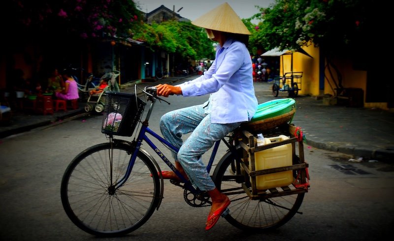 Local Vietnamese woman wearing a traditional conical hat and blue long-sleeved shirt riding a bicycle through the quiet old streets of Hoi An, Vietnam, with baskets and plastic containers strapped to the back of her bike as she passes colorful houses and flowering trees in the ancient town
