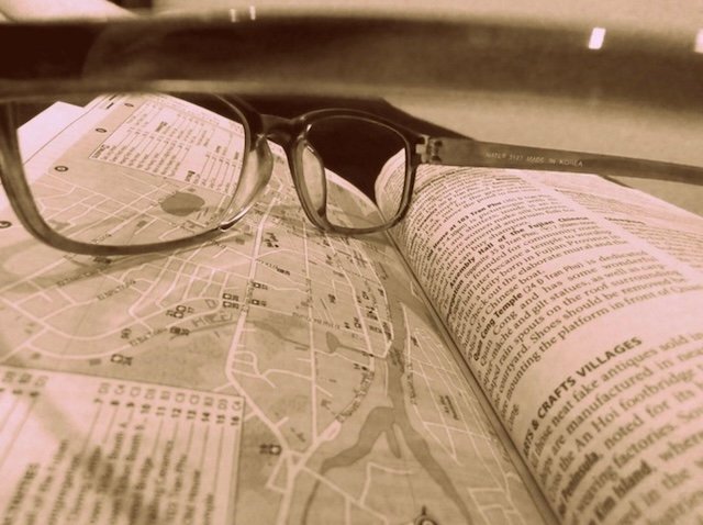 Sepia-toned close-up of reading glasses resting across an open Lonely Planet Vietnam guidebook showing a detailed map of Hoi An and surrounding text about local craft villages, symbolizing travel planning and preparing for a day of shopping for custom-made clothes in the historic Vietnamese town