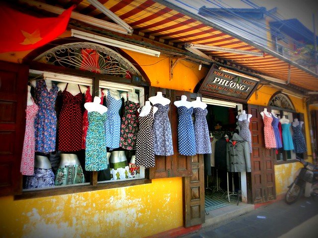 Bright and colorful tailor shop in Hoi An, Vietnam, with rows of custom-made dresses in floral and polka dot patterns displayed on mannequins outside a yellow-painted storefront beneath a striped awning, showcasing the city’s famous tradition of bespoke clothing and handmade fashion