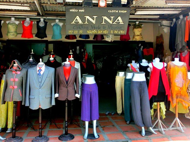 Tailor shop in Hoi An, Vietnam, displaying a variety of custom-made garments on mannequins including tailored suits, blazers, skirts, trousers, and vibrant traditional-inspired tops, with additional clothing hanging inside the boutique under a sign reading “An Na Cloth Shop” along Nguyen Duy Hieu Street