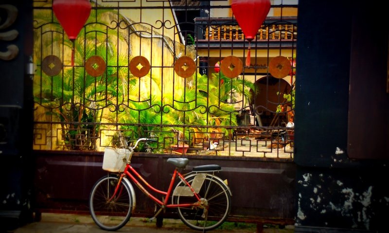 A vintage red bicycle with a front basket parked against an ornate wrought-iron gate decorated with circular motifs and flanked by red lanterns in the historic town of Hoi An, Vietnam, with lush green palms and a yellow-painted courtyard visible in the background