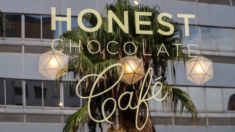 Elegant glass storefront sign of Honest Chocolate Café in Cape Town, reflecting a palm tree and city buildings, inviting visitors to enjoy handcrafted bean-to-bar chocolate treats