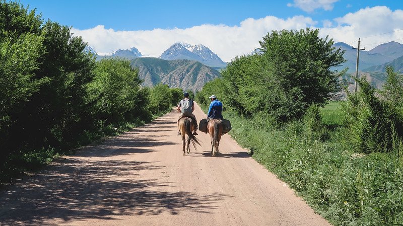 Two horse riders beginning the 3-Day Panorama Trek in Kyrgyzstan, riding along a dirt road lined with lush greenery towards the distant snow-capped Tian Shan Mountains under a bright blue sky