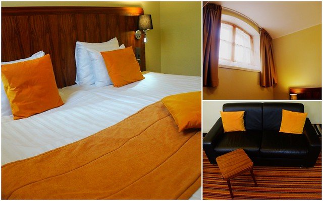 Hotel Katajanokka room in Helsinki, Finland, featuring a comfortable double bed with orange pillows and blanket, arched window with curtains, and a black leather sofa, blending cozy modern design with the historic character of the former jailhouse
