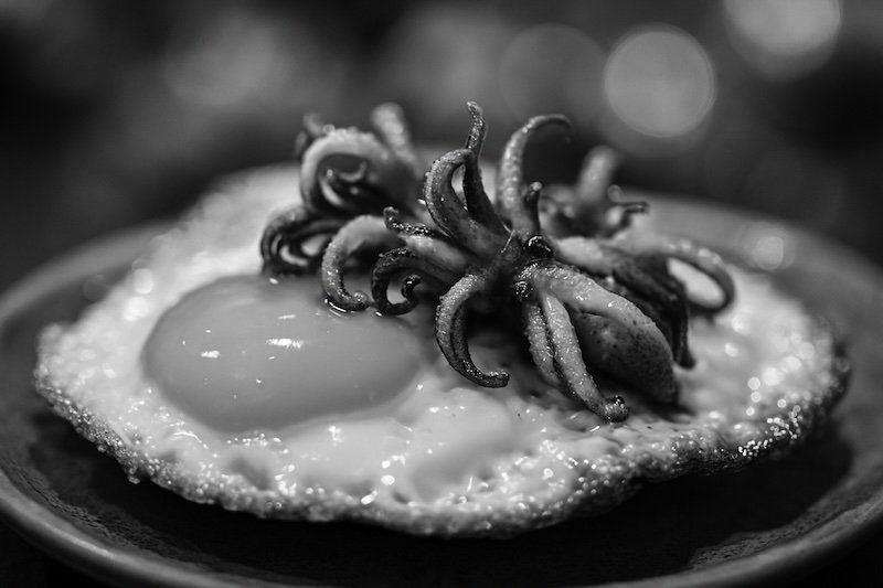 A black and white macro photograph of huevos con chipirones served at Barcelona’s La Boqueria Market. The image focuses on the glossy egg yolk, crispy edges, and tender baby squid with curled tentacles, set against a soft bokeh background that emphasizes texture and depth A black and white macro photograph of huevos con chipirones served at Barcelona’s La Boqueria Market. The image focuses on the glossy egg yolk, crispy edges, and tender baby squid with curled tentacles, set against a soft bokeh background that emphasizes texture and depth