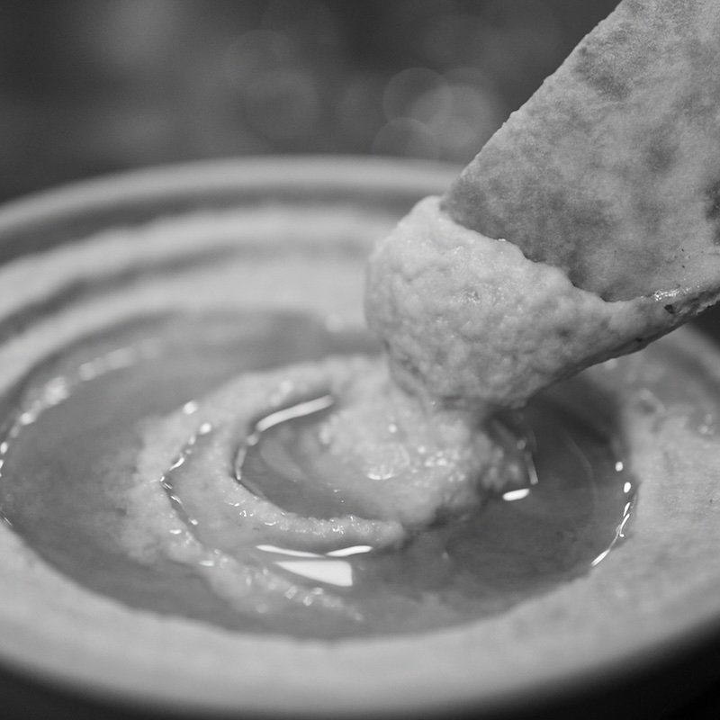 A black and white macro photograph of pita bread being dipped into a bowl of hummus generously topped with olive oil. The image focuses on the smooth hummus and glistening oil textures, with strong bokeh in the background, creating a rich, appetizing close-up