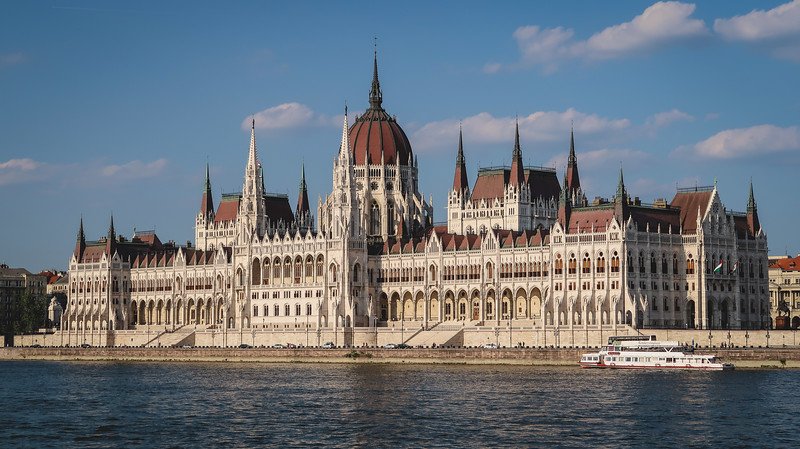 The majestic Hungarian Parliament Building in Budapest viewed from across the Danube River on a sunny day, with a tour boat passing by. Touring this iconic Gothic Revival landmark is one of the top things to do when visiting Budapest, Hungary.