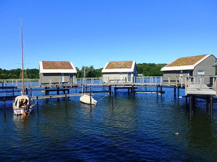 Modern overwater bungalows with green roofs at Im-Jaich Wasserferienwelt on Rügen Island, Germany, surrounded by calm marina waters and small sailboats under a clear blue summer sky