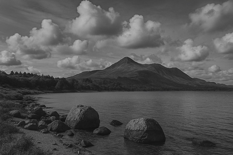 A black and white photograph of the Isle of Arran in Scotland, showing a tranquil shoreline with large boulders in the foreground, calm reflective water, and a striking mountain peak rising in the distance beneath a sky of dramatic clouds. The scene feels serene and timeless A black and white photograph of the Isle of Arran in Scotland, showing a tranquil shoreline with large boulders in the foreground, calm reflective water, and a striking mountain peak rising in the distance beneath a sky of dramatic clouds. The scene feels serene and timeless