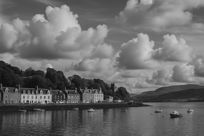 A black and white photograph of Tobermory Harbour on the Isle of Mull, Scotland, showing a row of charming waterfront buildings backed by trees and a church on a hill. Several small boats rest in the calm bay, with rolling hills and dramatic clouds completing the peaceful scene A black and white photograph of Tobermory Harbour on the Isle of Mull, Scotland, showing a row of charming waterfront buildings backed by trees and a church on a hill. Several small boats rest in the calm bay, with rolling hills and dramatic clouds completing the peaceful scene