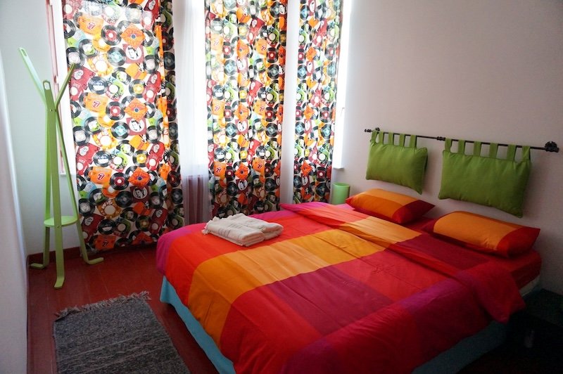 A colorful and modern Airbnb bedroom in Istanbul, Turkey, featuring a vibrant red, orange, and pink bedspread, playful vinyl record-print curtains, green wall cushions, and minimalist decor