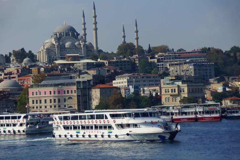 Passenger ferries crossing the Bosphorus Strait in Istanbul, Turkey, with the grand domes and towering minarets of the Süleymaniye Mosque rising above the city skyline in the background