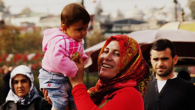 A joyful candid street scene in Istanbul, Turkey, showing a smiling woman in a colorful headscarf lifting a baby in a pink hoodie, surrounded by onlookers in a lively public square