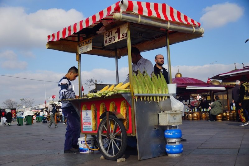 Street food vendor cart selling freshly roasted and boiled corn on the cob under a red-and-white striped canopy at an outdoor market square in Istanbul, Turkey, with locals and tourists passing by in the background