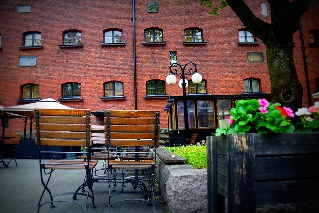 Jailyard Terrace at Hotel Katajanokka in Helsinki, Finland, featuring outdoor seating with wooden chairs, flower planters, and views of the historic red-brick former prison building, now transformed into a stylish hotel with unique dining spaces