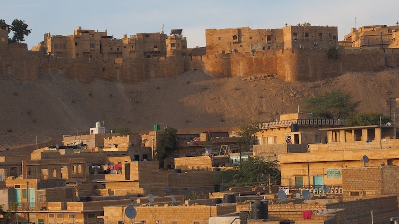 Panoramic view of Jaisalmer Fort rising above the golden sandstone rooftops of Jaisalmer, Rajasthan, India, captured from a distant rooftop vantage point at sunset