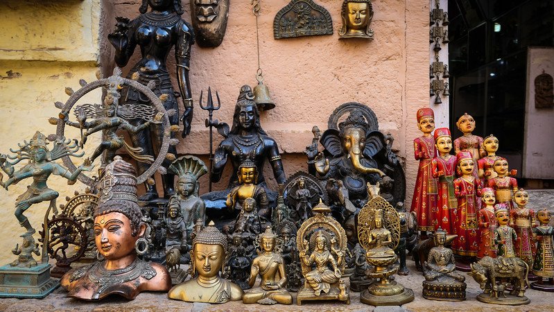 Colorful souvenir shop display in Jaisalmer, India, featuring intricately crafted brass statues of Hindu deities, Buddha figures, and traditional Rajasthani wooden dolls arranged on a stone ledge