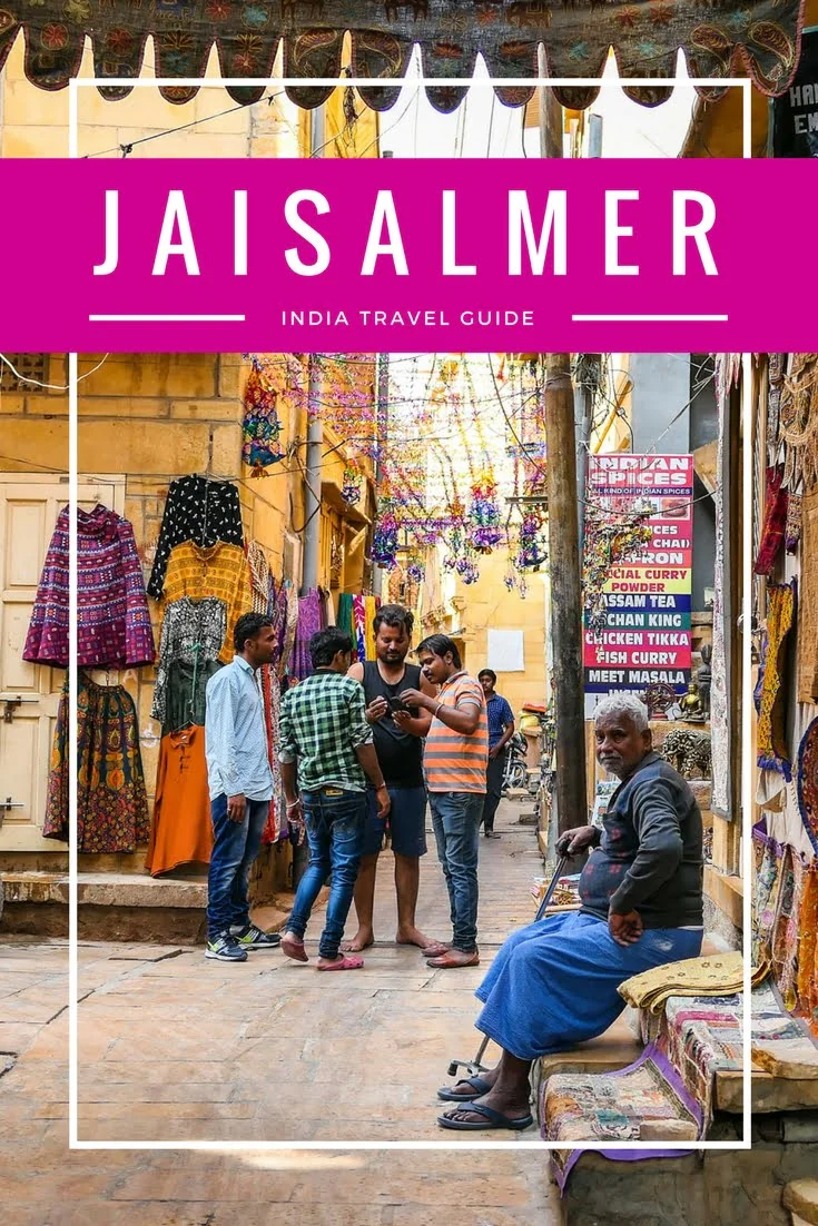 Colorful market alley in Jaisalmer, India, with locals chatting under strings of hanging decorations and vibrant textiles on display, featured in a Jaisalmer travel guide to Thar Desert adventures