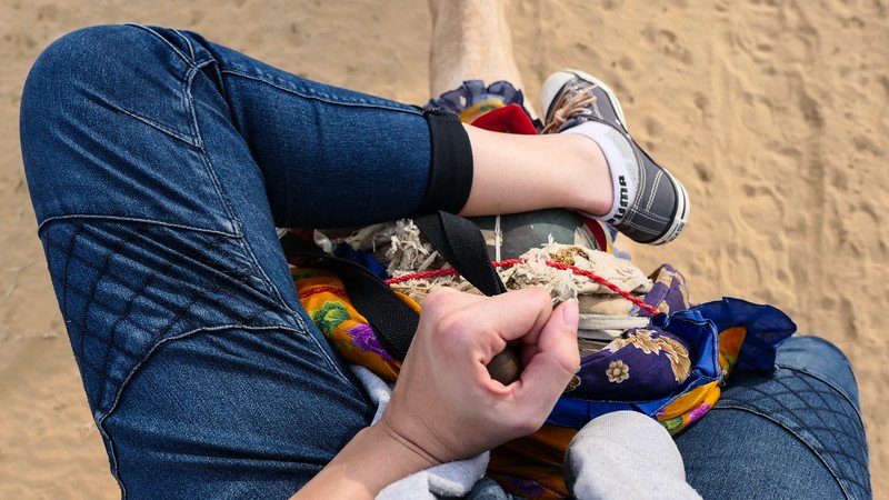 Point-of-view shot of Audrey Bergner from That Backpacker riding a camel on a desert safari near Jaisalmer, India, showing her legs, casual sneakers, and the camel’s colorful saddle blankets against the sandy Thar Desert backdrop