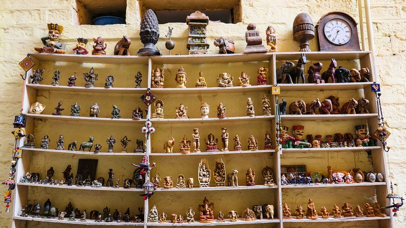 Display shelves in Jaisalmer, India, filled with traditional Rajasthani souvenirs including miniature statues, carved figurines, wooden elephants, deities, and handcrafted trinkets inside a sandstone shop
