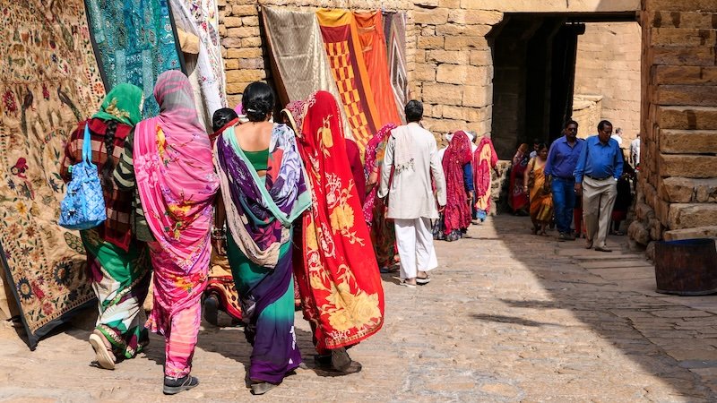 Group of women wearing vibrant, colorful saris walking through a sunlit stone alley lined with hanging textiles in Jaisalmer, Rajasthan, India, capturing the region’s rich traditional attire and lively market atmosphere