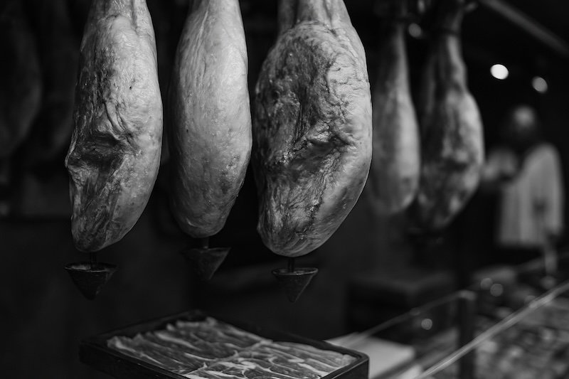 A black and white photograph of Jamón Serrano hanging in a traditional stall at Barcelona’s La Boqueria Market. The image focuses on the textured surface and marbled fat of the cured hams, with soft bokeh in the background adding depth and atmosphere A black and white photograph of Jamón Serrano hanging in a traditional stall at Barcelona’s La Boqueria Market. The image focuses on the textured surface and marbled fat of the cured hams, with soft bokeh in the background adding depth and atmosphere