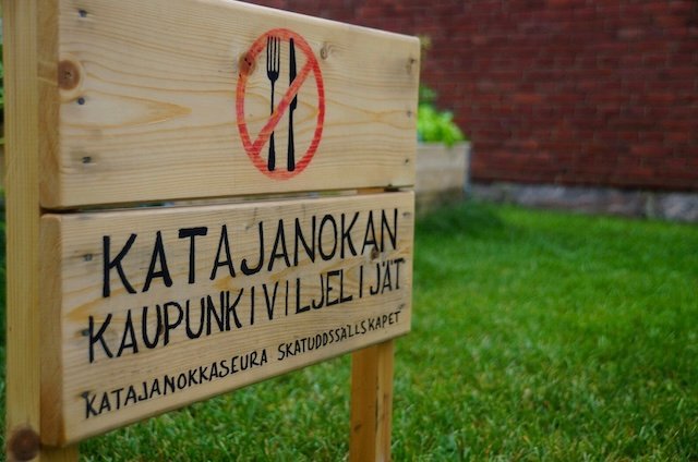 Wooden signboard in Finnish reading “Katajanokan Kaupunkiviljelijät” placed on a grassy lawn in Helsinki, Finland, near Hotel Katajanokka, highlighting local community gardening culture beside the historic red-brick former prison building
