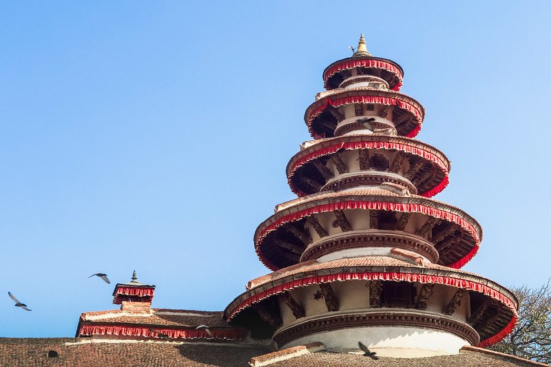Multi-tiered pagoda-style temple in Kathmandu Durbar Square, Nepal, with intricate wood carvings, red-trimmed roofs, and birds flying against a clear blue sky