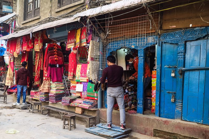 Street market in Kathmandu, Nepal with vibrant fabrics, traditional clothing, and shoe stalls set against colorful blue doors and bustling local shoppers