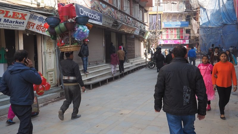 Street scene in Kathmandu with a local man balancing baskets of goods on his head, surrounded by pedestrians and daily market life in Nepal