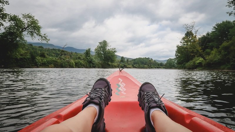 Relaxing solo kayaking adventure on the Missisquoi River with feet propped on a red kayak, surrounded by lush green hills in Sutton, Quebec’s Eastern Townships under overcast skies