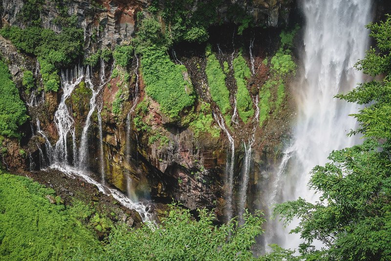 Close-up view of the rugged cliffside at Kegon Falls in Nikko, Japan, showing delicate side streams trickling down moss-covered rock walls beside the thundering main waterfall
