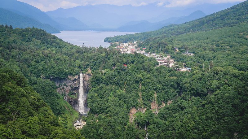 Scenic view from the Akechidaira Ropeway Observation Point overlooking the dramatic Kegon Falls cascading down a cliff into a lush green gorge, with the tranquil waters of Lake Chuzenji and the mountain village of Nikko, Japan, framed by misty blue peaks in the distance