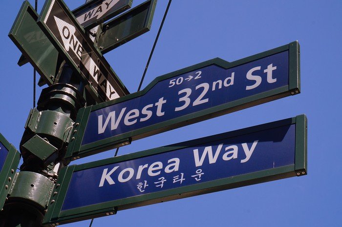 Close-up of intersecting street signs for West 32nd Street and Korea Way in New York City’s Koreatown, set against a clear blue sky. This vibrant stretch of Midtown Manhattan is famous for its Korean restaurants, bakeries, and late-night culture