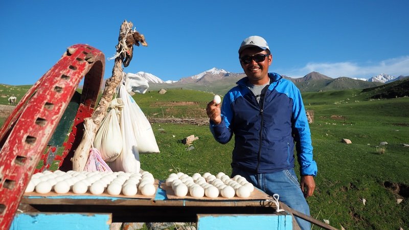 Our local guide in Kyrgyzstan smiling while holding a traditional dried dairy snack (kurut) beside a table full of round white buns, with green pastures and snow-capped mountains in the background during the 3-Day Panorama Trek
