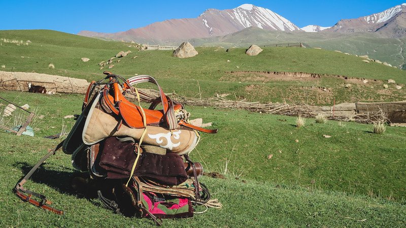 A colorful traditional Kyrgyz horse saddle resting on the grass with snow-capped mountains and rolling green hills in the background during the 3-Day Panorama Trek in Kyrgyzstan