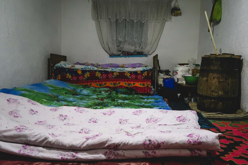 A cozy homestay bedroom in rural Kyrgyzstan with colorful patterned bedding, floor mattresses, a lace-curtained window, and traditional household items during the 3-Day Panorama Trek
