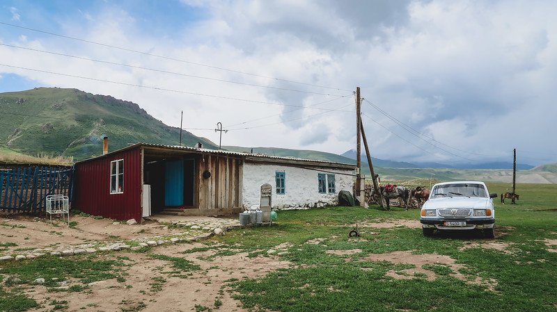 A rustic countryside homestay kitchen and dining building in rural Kyrgyzstan, with a vintage white car parked out front, horses tied nearby, and green hills and dramatic clouds in the background during the 3-Day Panorama Trek
