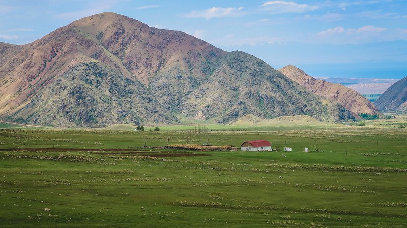 A small homestay with a red roof nestled in a wide green valley surrounded by rugged mountains in Kyrgyzstan, viewed from a hill during the 3-Day Panorama Trek