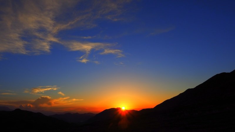 A brilliant sunset over the mountains in Kyrgyzstan, with the sun dipping behind dark silhouetted peaks and the sky glowing in rich orange, red, and deep blue during the 3-Day Panorama Trek